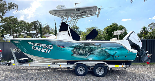 A white and teal custom fishing boat wrap named 'Popping Candy' with a large Giant Trevally graphic on the side, mounted on a dual-axle trailer outdoors.