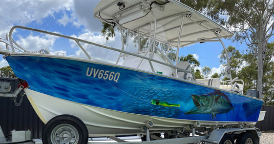 Blue and white boat featuring a detailed Wahoo fish boat wrap, parked on a trailer under a blue sky with clouds.